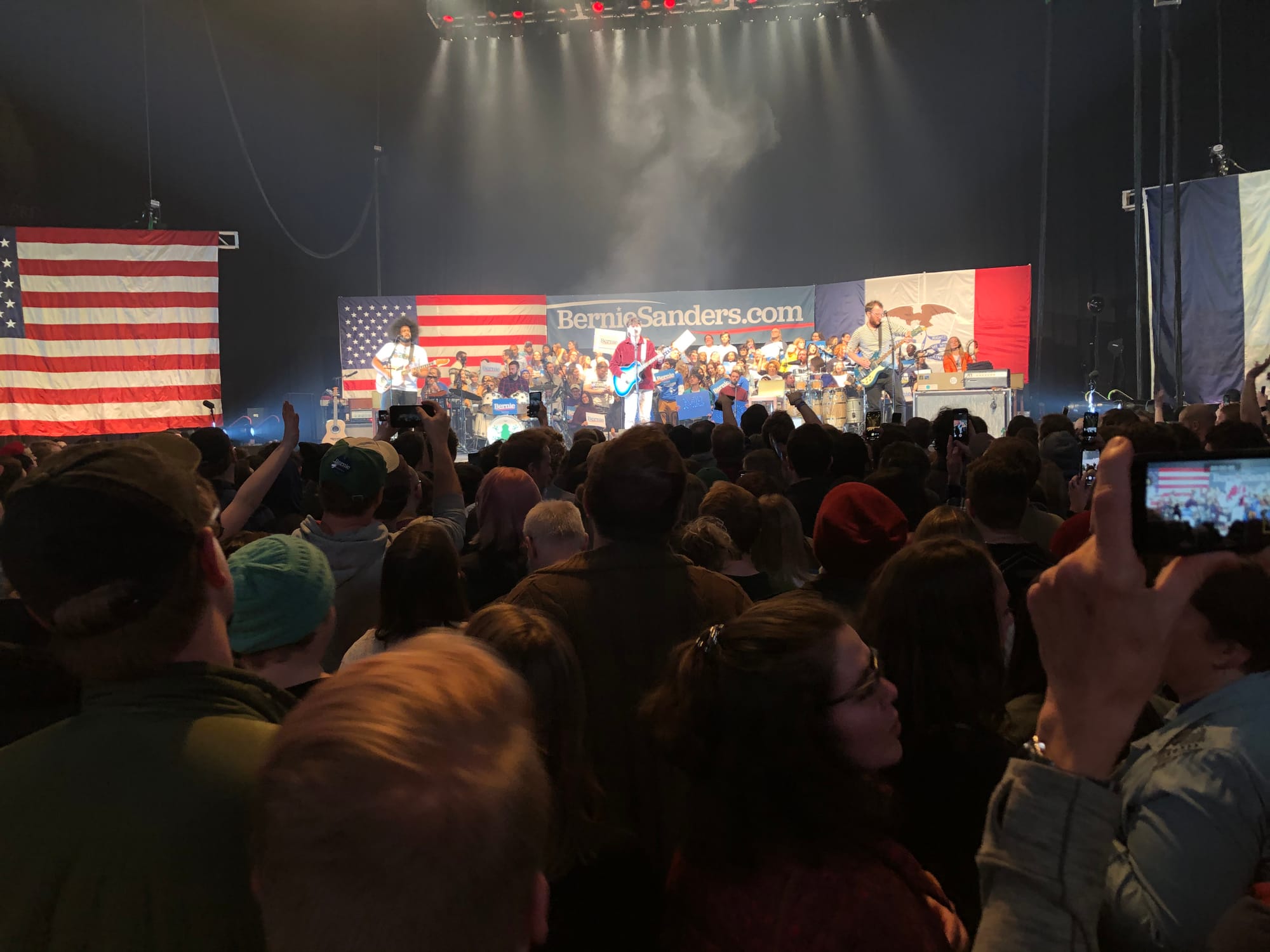 Ezra Koenig strums the guitar on large stage. A crowd of 50 plus people sit on the risers behind him. Dozens of people stand on the ground facing him. Above Ezra there is an American Flag, an Iowa state flag, and a sign reads: BernieSanders.com. 