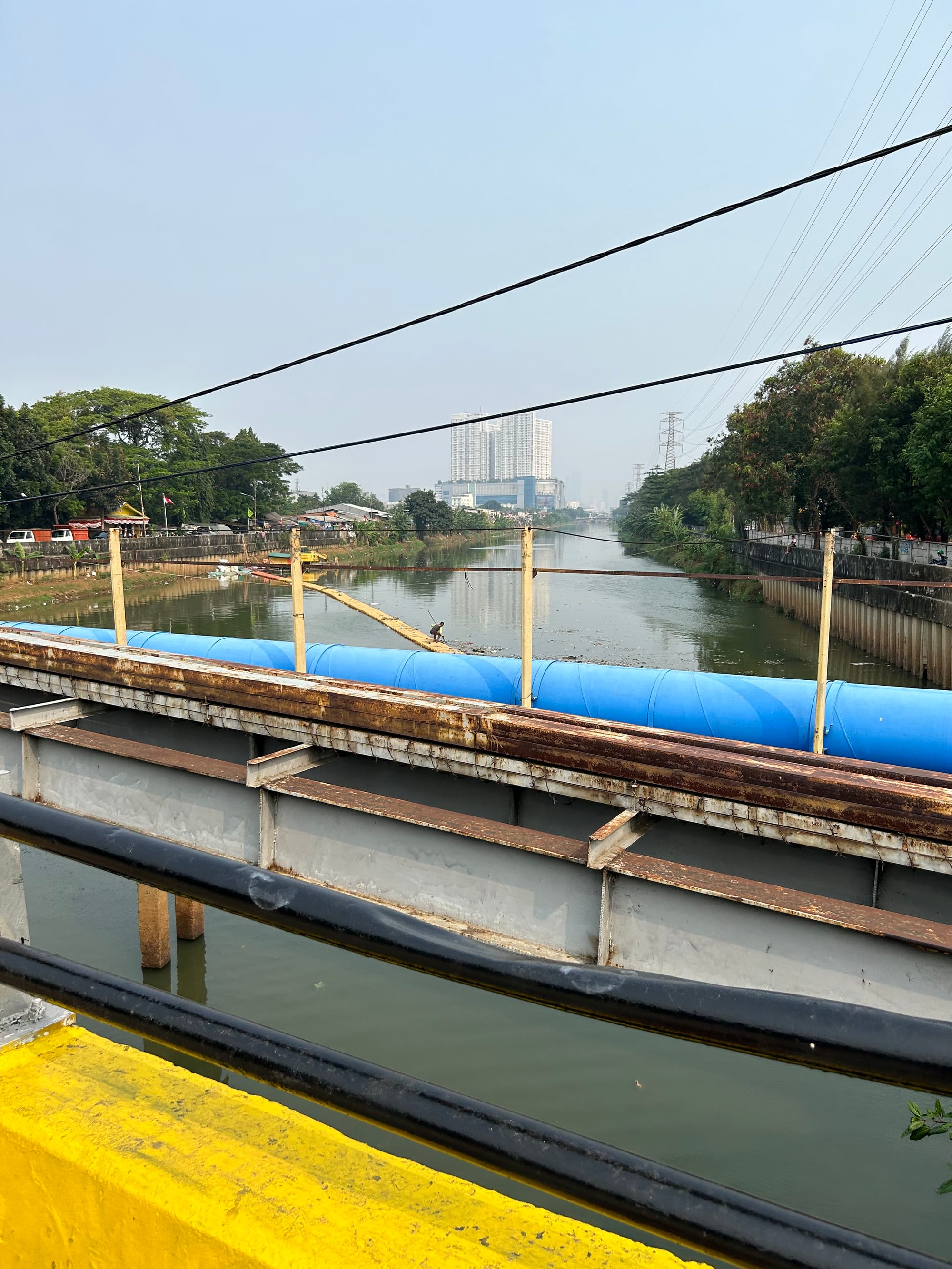 Jakarta: view from a rusty steel bridge with vivid yellow painted concrete walls. Beneath in the distance, a man on a narrow pontoon bridge holding a spear or net