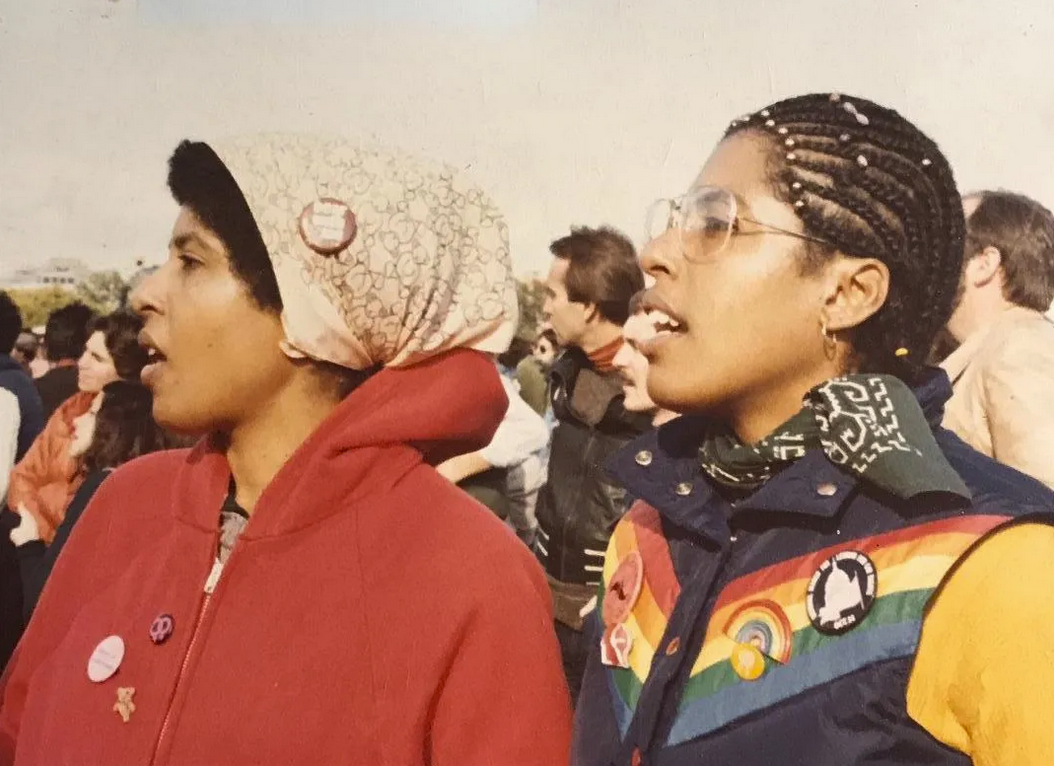 Two young Black women at a 1979 march for gay rights in Washington, in colorful gear, protest buttons and bandannas, seemingly absorbed in a speech being given offscreen 