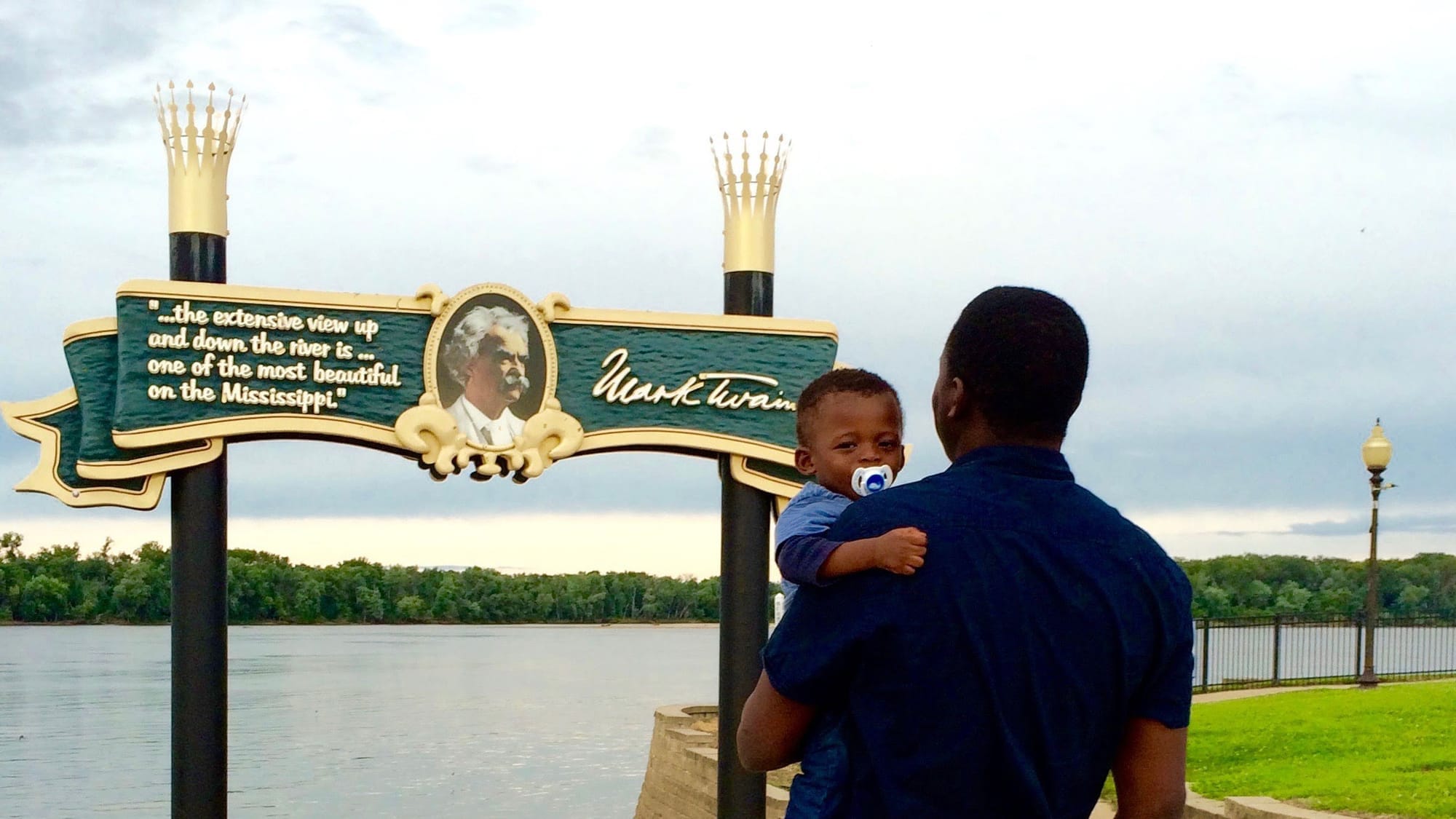 The author and his baby son in Hannibal, Missouri, standing before the Mississippi River, and a large, elaborate sign depicting Mark Twain, with the quote: "... the extensive view up and down the river is... one of the most beautiful on the Mississippi."
