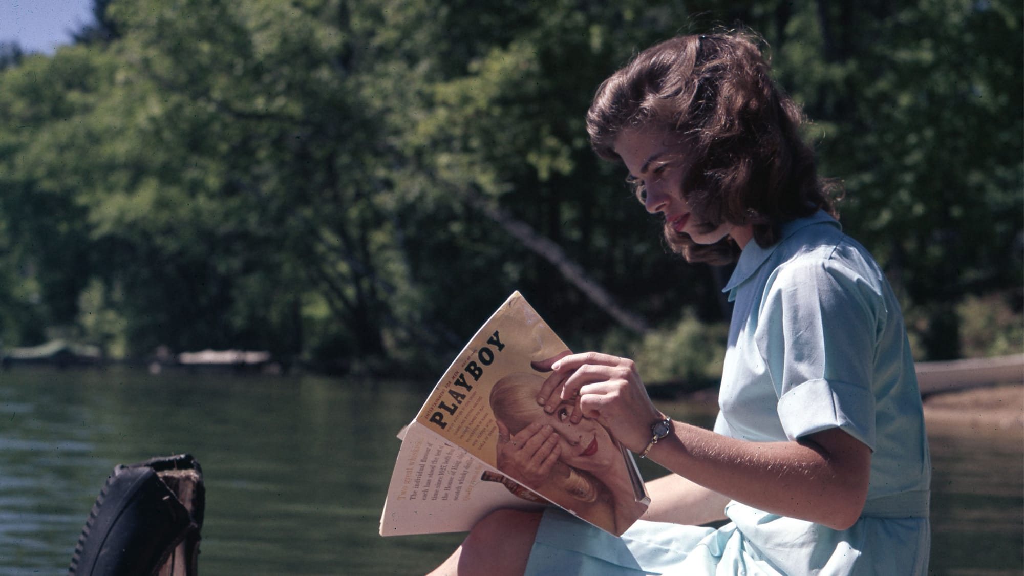A young woman in 60s shirt and blouse of crisp pale blue cotton, seated lakeside on a wooden pier, reads Playboy Magazine