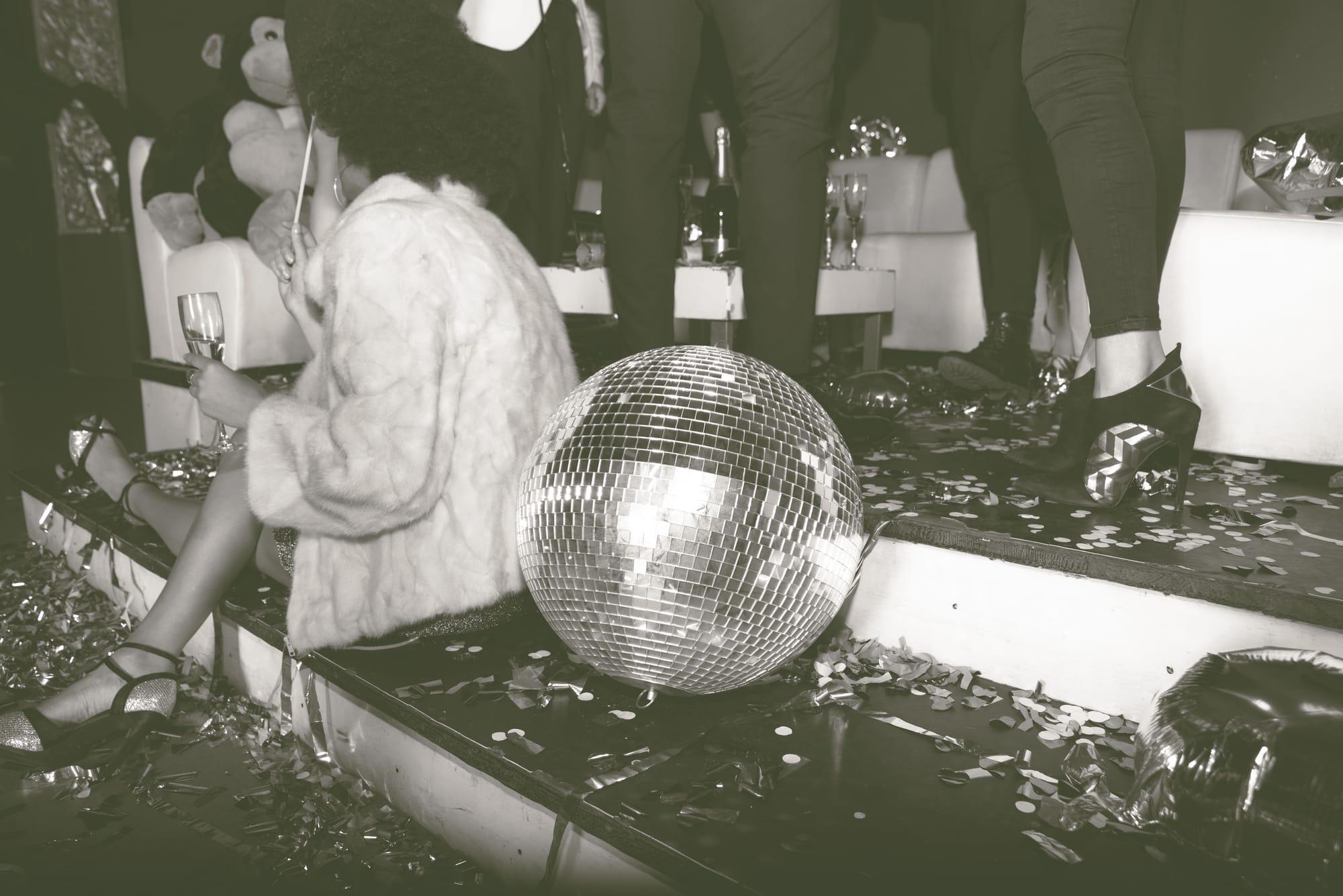 A lady in black satin heels and a white plush jacket seated next to a mirror ball on the steps of a nightclub; her back is to us ad she's holding a champagne flute. Behind her we can see a big stuffed chimp on a long white banquette, a bottle of champagne and more flutes on a low table, and the booted legs and high heels of fellow partygoers