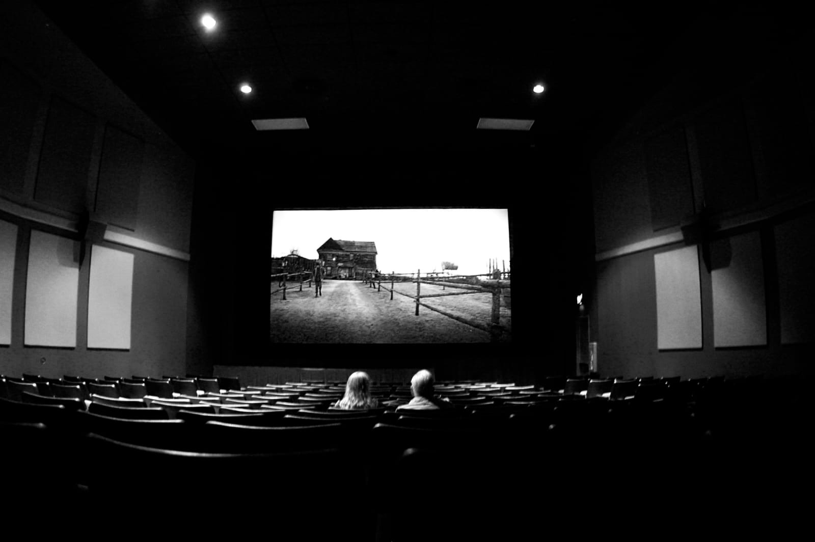 black and white image of a darkened movie theater with just two patrons watching what appears to be a bleak Western scene