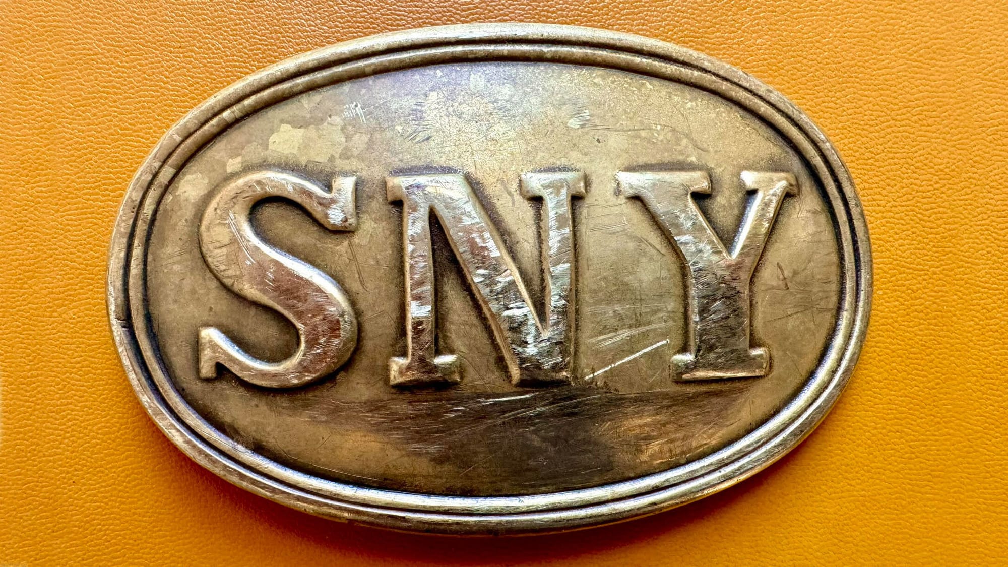 shiny belt buckle with an antique finish reading S N Y against a leather background