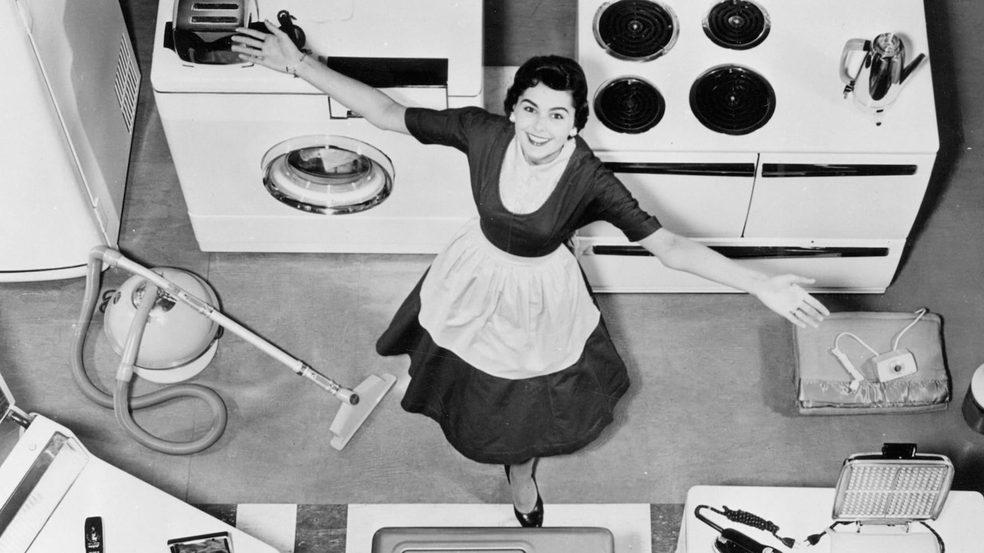 B/W image from the early 50's shows a woman in a black New Look dress and white apron in a spotless kitchen