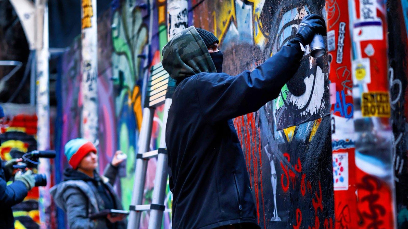 The artist Hugus in a camouflage hood, dark jacket and black gloves, at work on a colorful mural