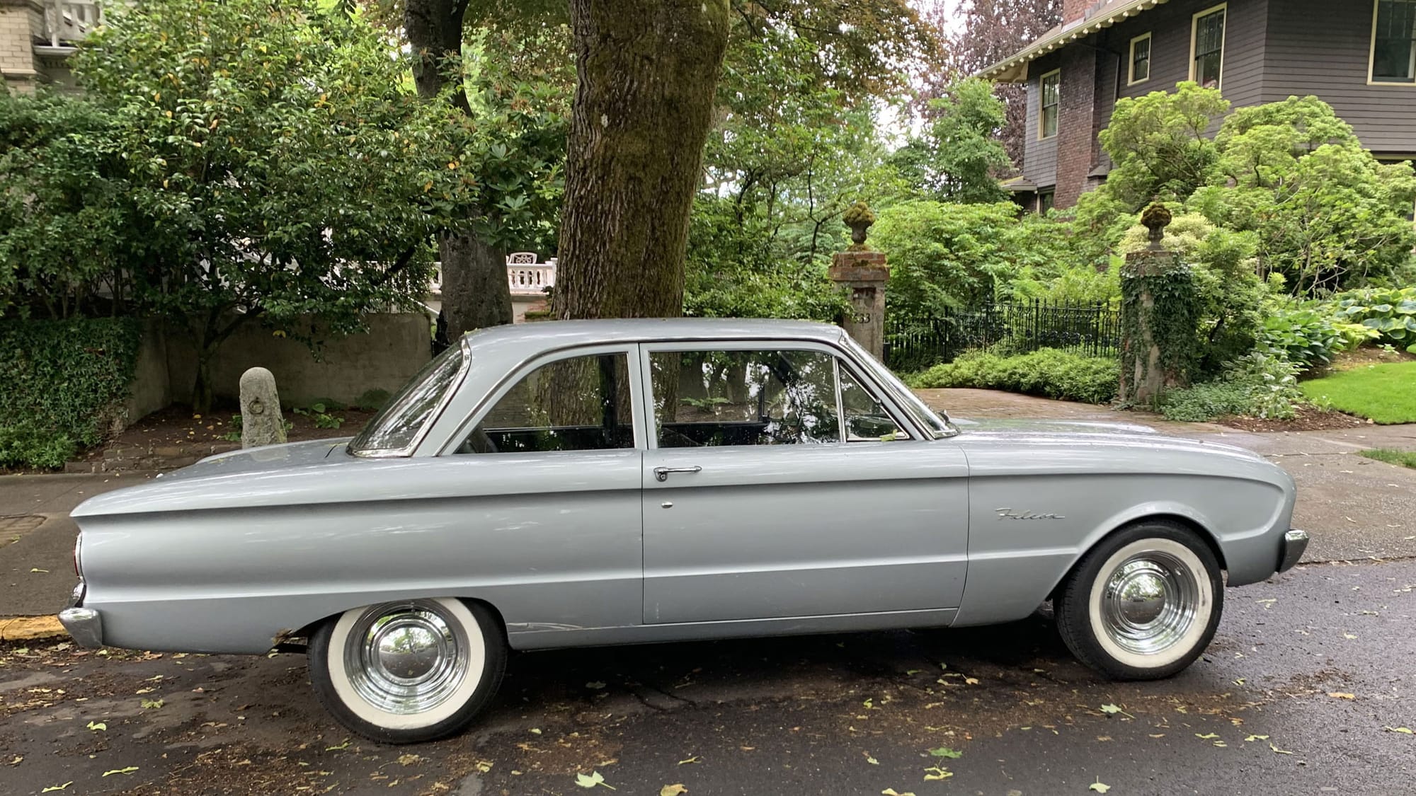 A silver Ford Falcon coupe parked in a handsome, leafy residential area, before a stone and wrought-iron gate 