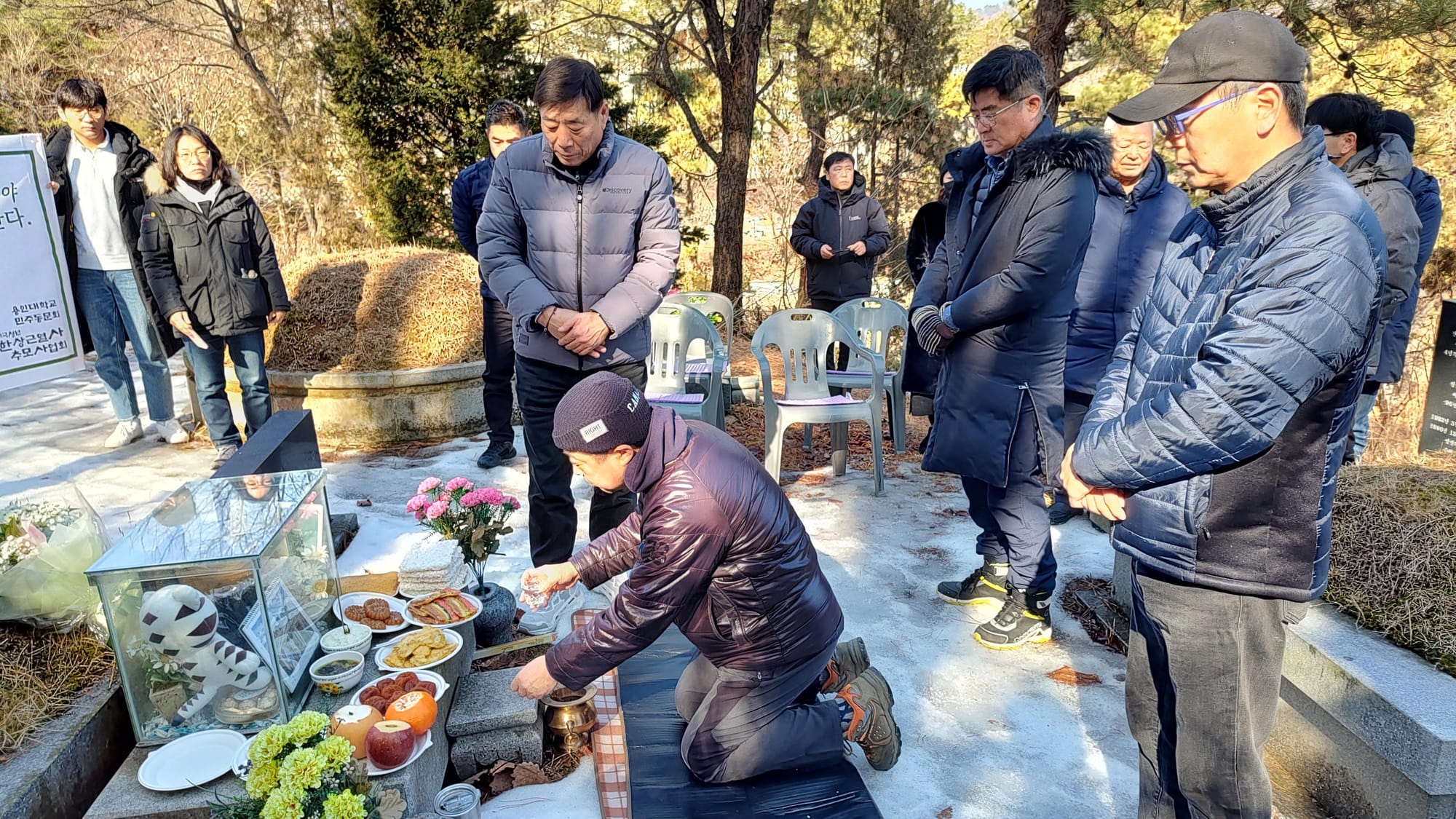 Memorial service for the 27th anniversary of the death of Han Sang-geun at the National Democratic Martyrs' Cemetery in Moran Park, Maseok. The image shows a number of winter coat clad people stand around a shrine on a bright day. There are fruits, cookies and other delicious foods on the altar, vases full of pink and yellow flowers, and a clear box with a cat plushie inside.