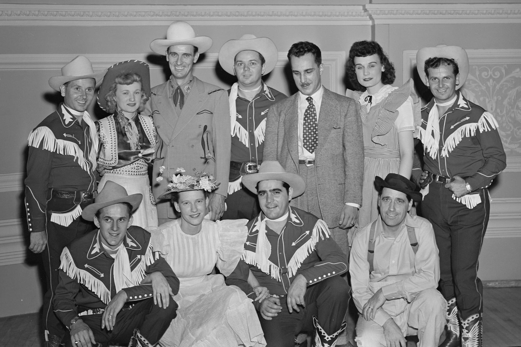 Black and white photo shows eleven country music stars of 1947, all in fancy cowboy and cowgirl regalia, including fringed shirts, kerchiefs and intricately tooled leather boots; Minnie Pearl in flowered hat and shirtwaist among them