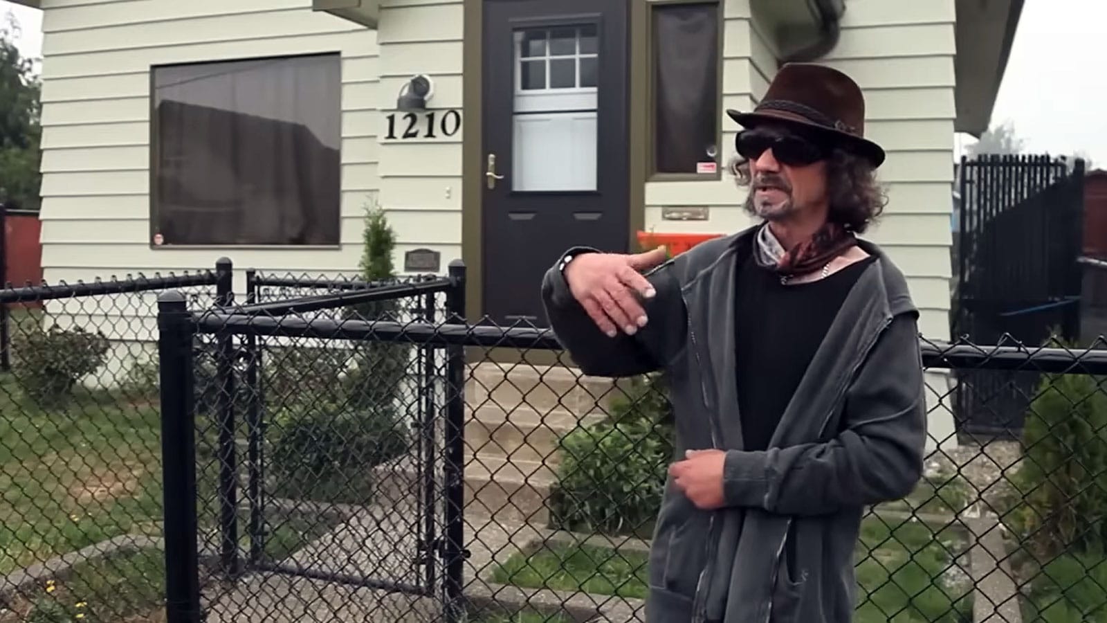 Nirvana's first drummer, Aaron Burckhard, in dark glasses and fedora, standing before a small house with chain-link fence