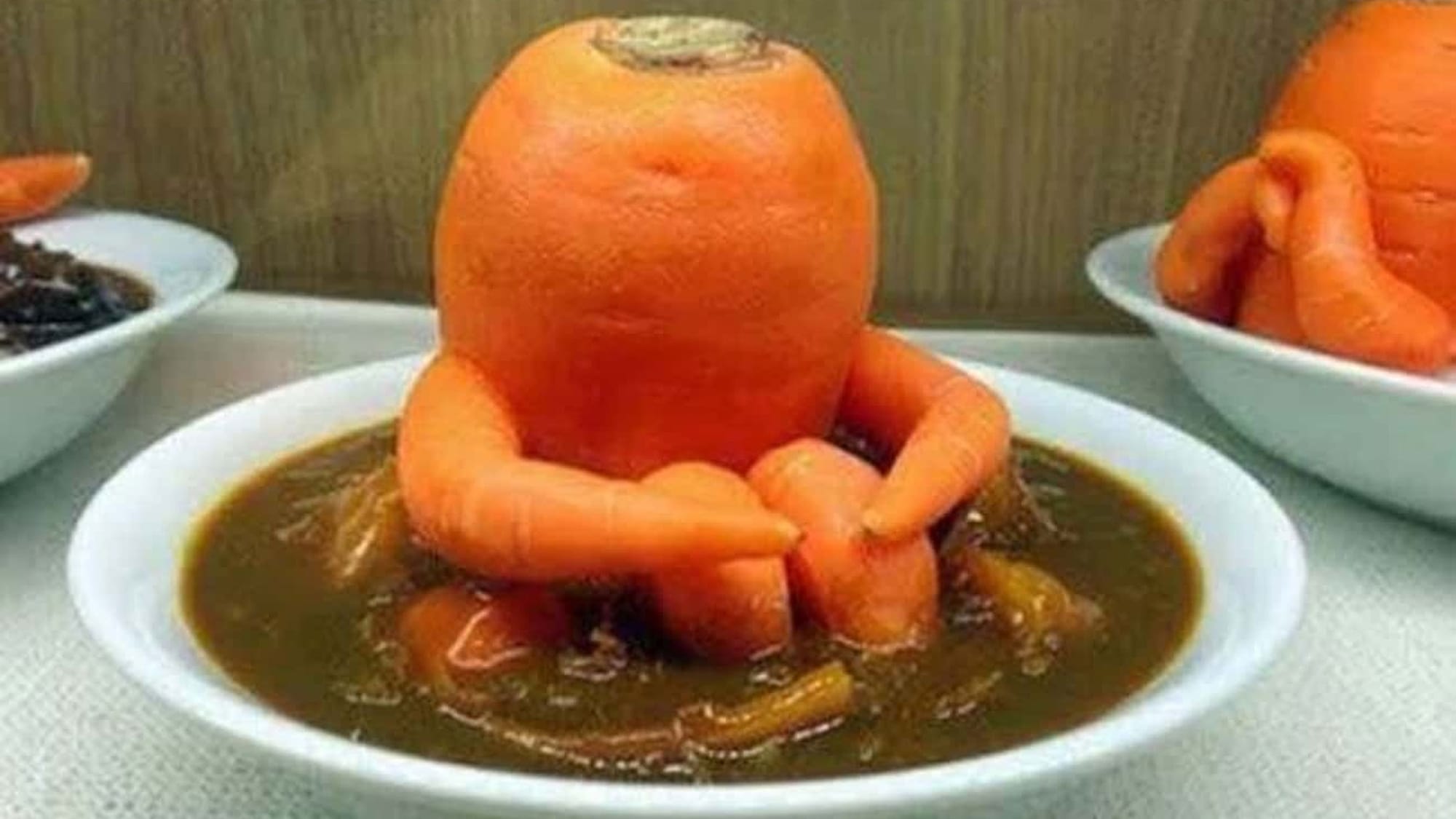 Meme image of a carrot sculpture resembling a person, comfortably seated, with carrot arms wrapped around carrot knees, in a bowl of soup