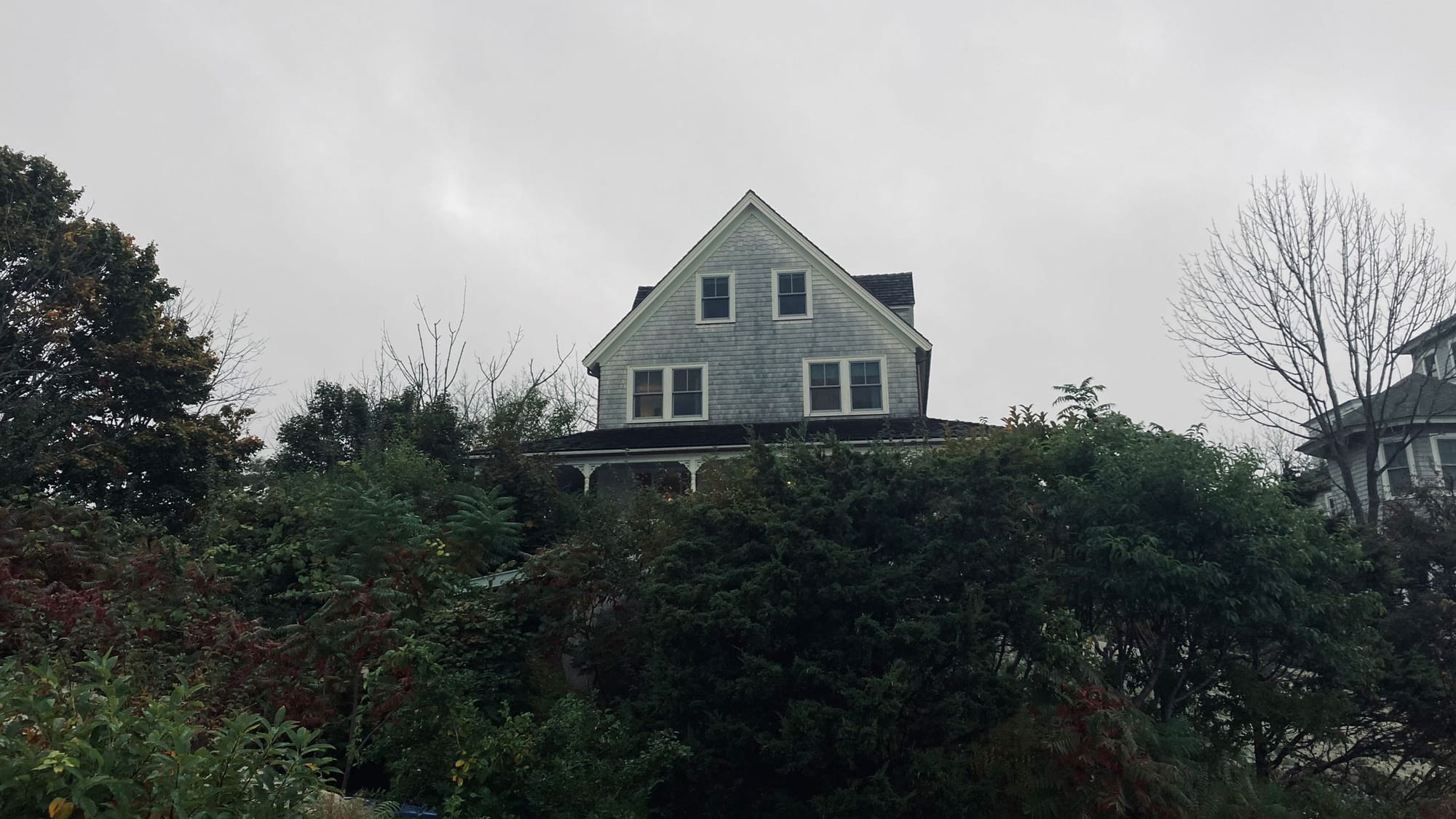 A wooden house stands above a green hillside near the sea in New England on a cloudy day