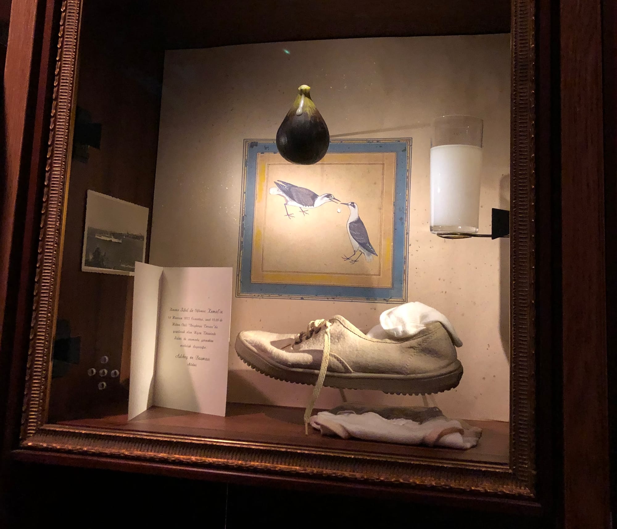 a shadow box holds a single worn white sneaker, a holiday card, glass of cloudy white raki, and a fig hanging from the top. A small illustration of two seagulls hangs in the back.