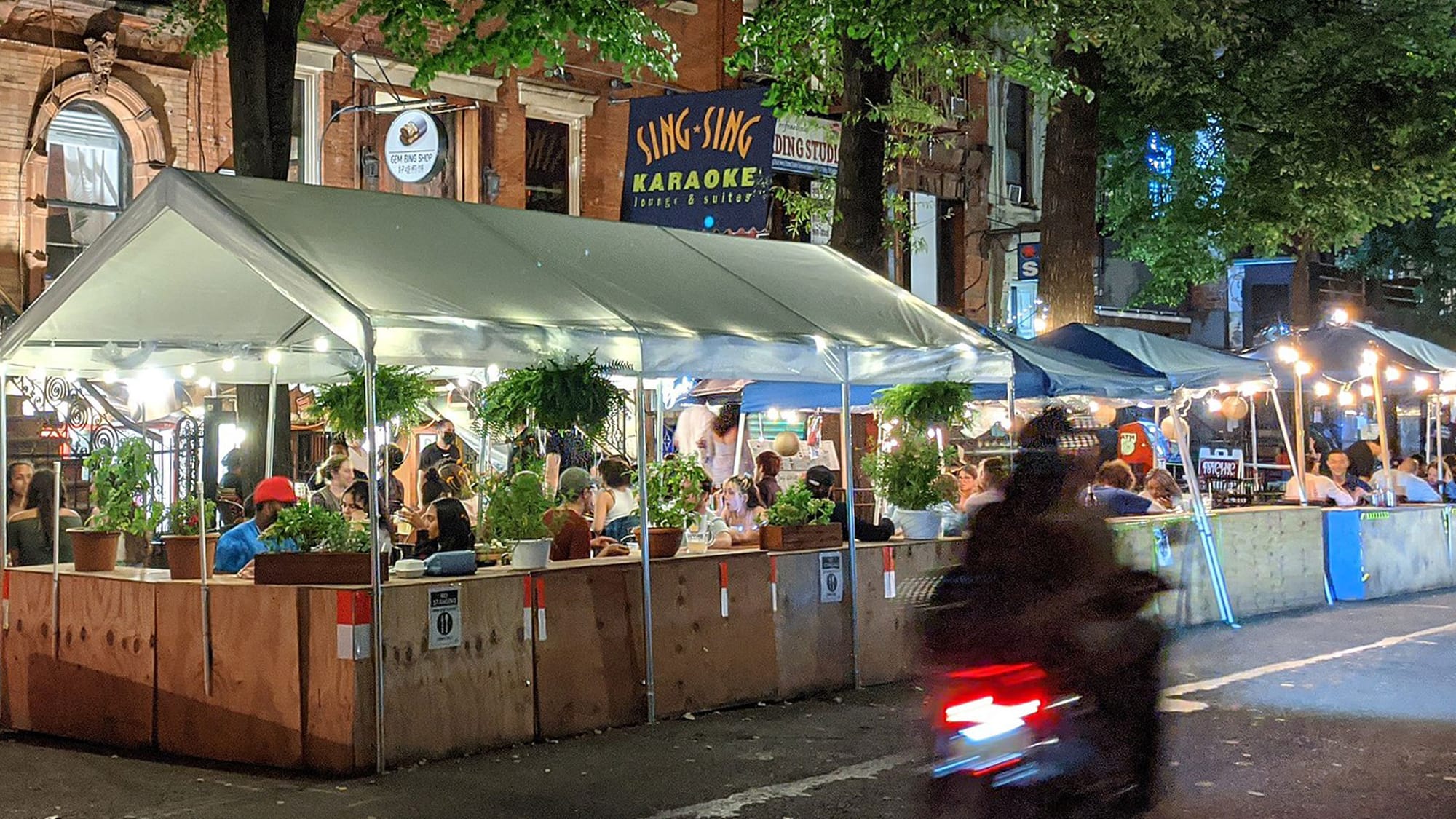 An outdoor dining setup on a tree-lined street outside the Sing-Sing Karaoke Lounge on St. Mark's in New York City's East Village during the pandemic, with lots of people eating, drinking and having fun, and a motorbike speeding past in the foreground