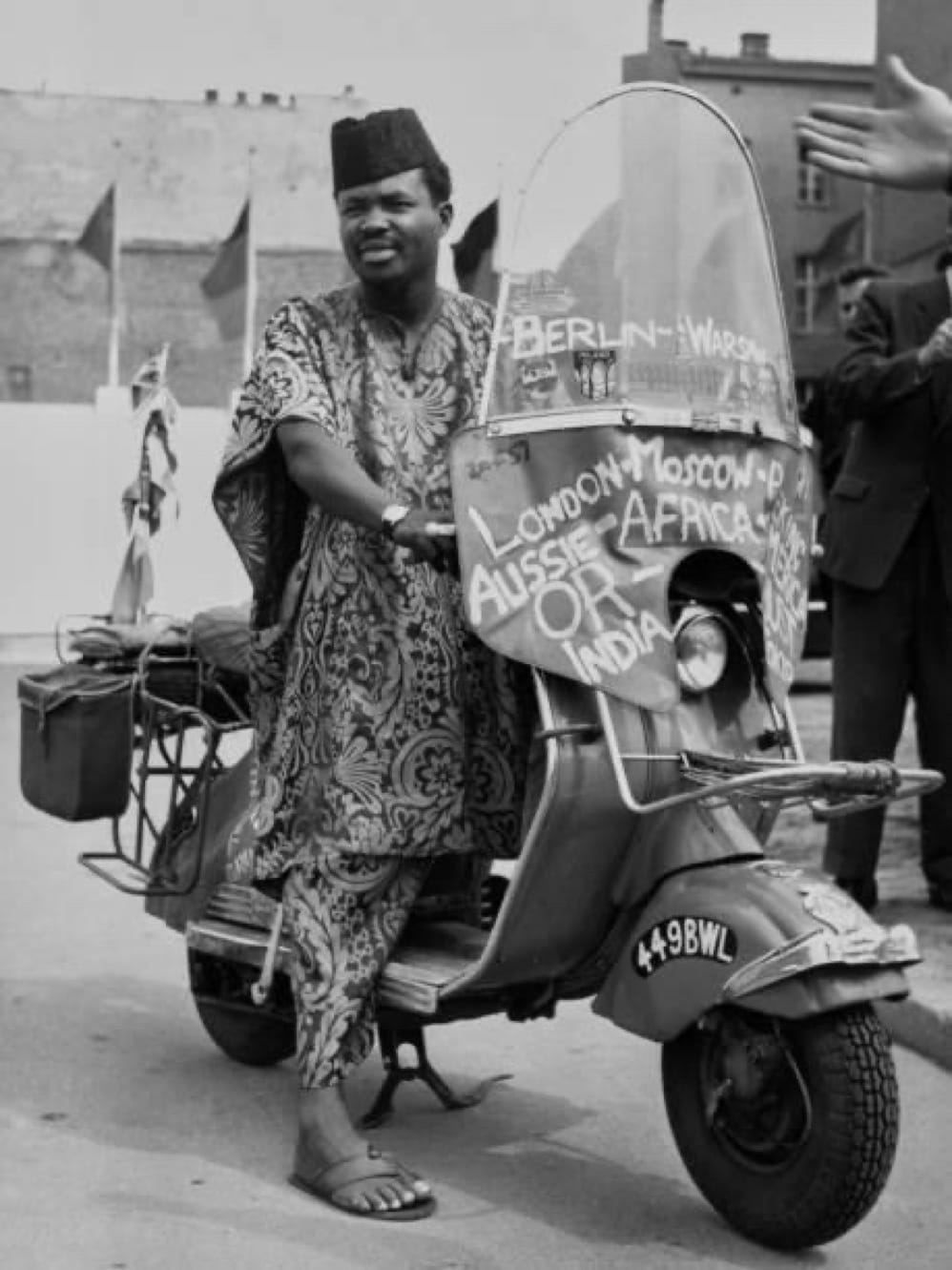Black and white photo of Ọlábísí Àjàlá posing on his scooter; he wears a traditional African smock in an intricate print, a dark cap, and leather sandals