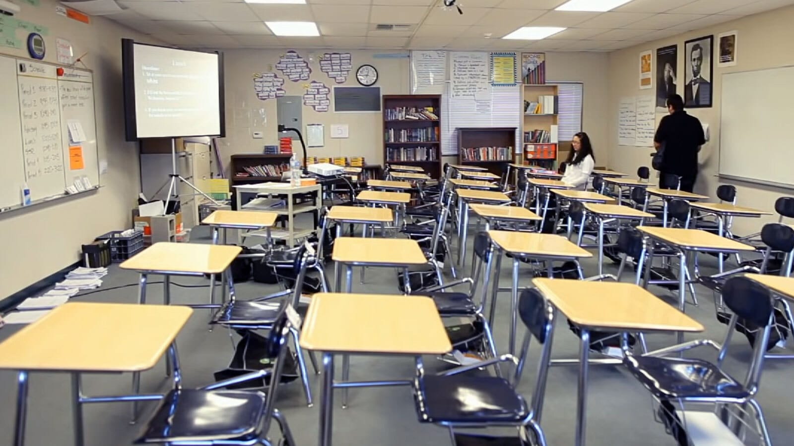 A classroom with five rows of desks, the kind that have black metal seats attached. Two people hang out in the back of the room, next to some bookshelves, posters and a poster of Abe Lincoln.
