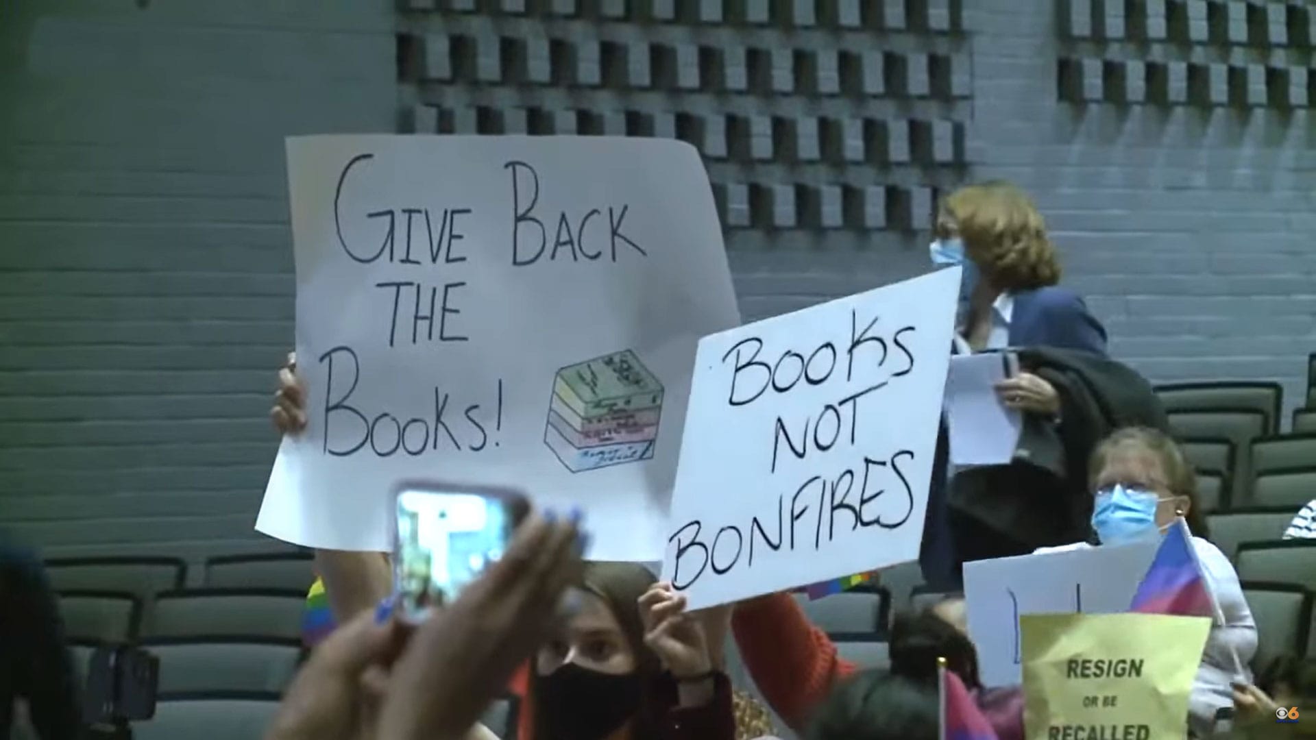 Screenshot from a news clip of a public hearing on book banning in Virginia: signs read "Books Not Bonfires" and "Give Back The Books!"