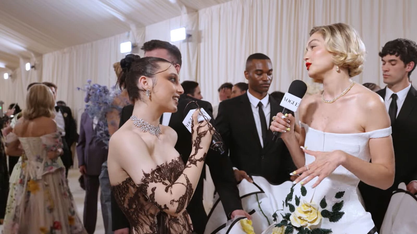 Emma Chamberlain, in a brown lace gown, tilts her head and smiles at Gigi Hadid, in an elaborate white ballgown embroidered with yellow flowers and green foliage; both are holding microphones