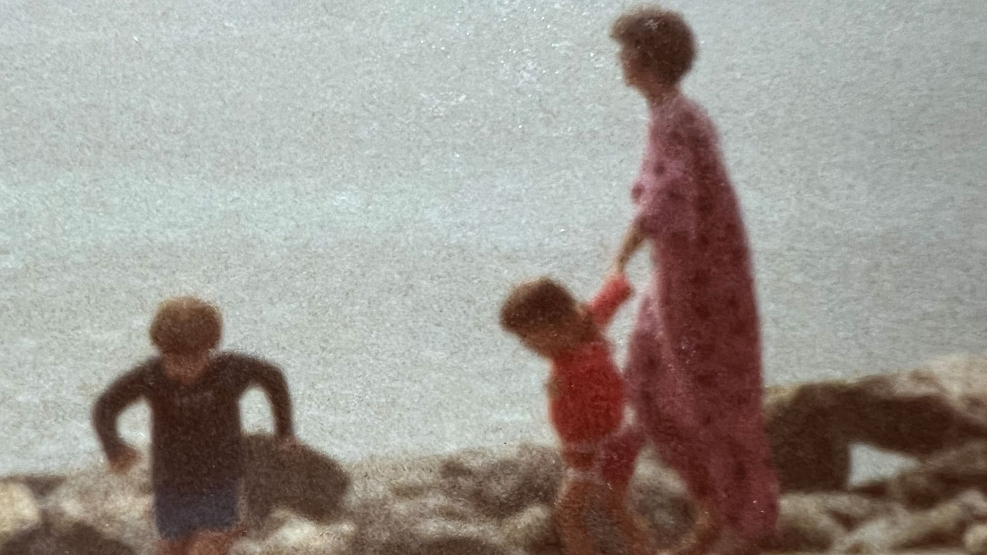 A powerfully romantic, blurry old family photo of a woman in a long pink beach caftan or robe with two little kids on a rocky shore, the palely glittering sea behind them