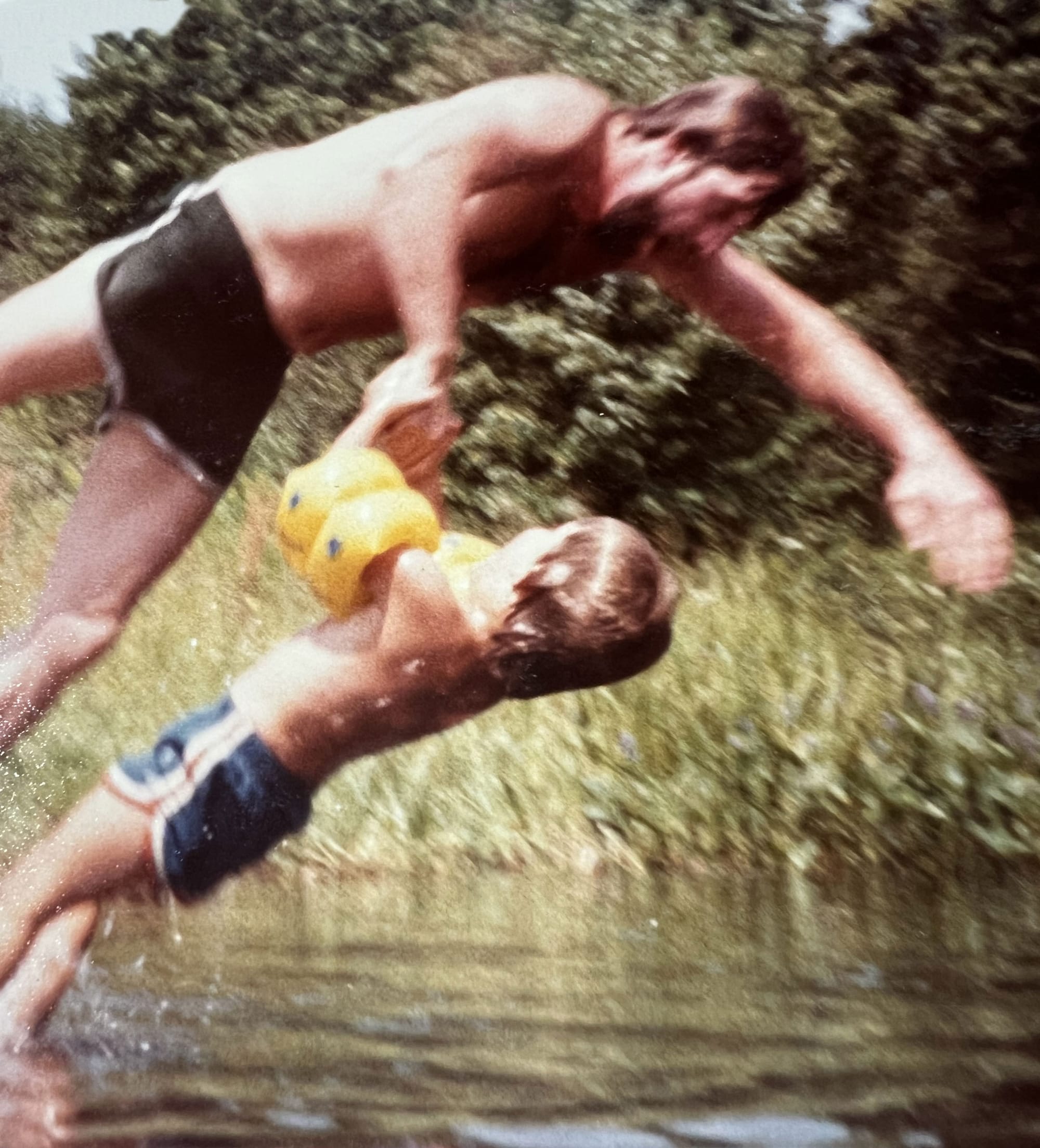 Father and small son, already soaking wet, in vintage trunks (the son in yellow water wings) diving and/or falling into a pond