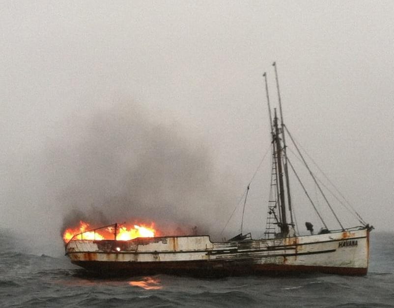 A fishing boat on the water, on fire, under a cloudy sky