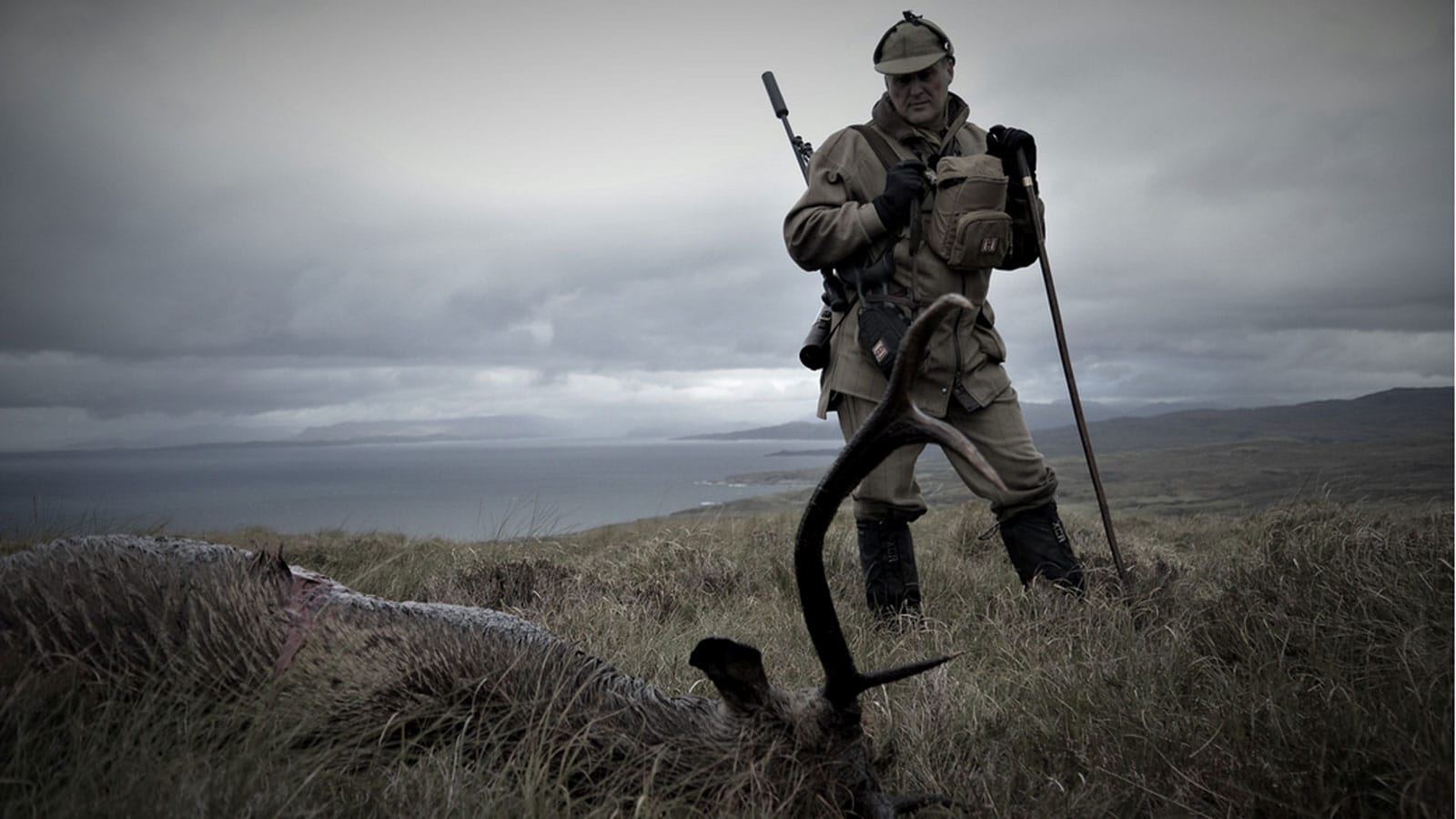 A deerstalker fully equipped for the hunt stands over the body of a buck, in a field by the sea under a cloudy sky