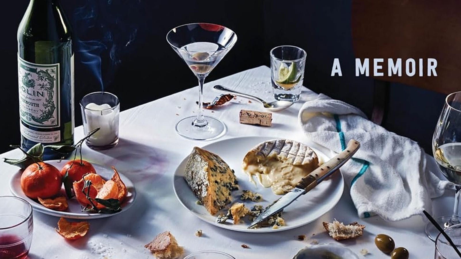 A table covered with a snow-white cloth bears the remains of a feast, with near-empty glasses, crusts of bread, cutlery, a cheese plate, olives, and fruit