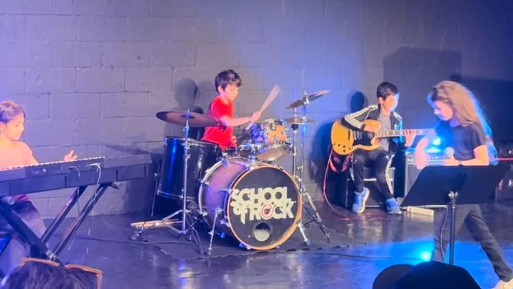 A small drummer in a red t-shirt performing with a 'School of Rock' band