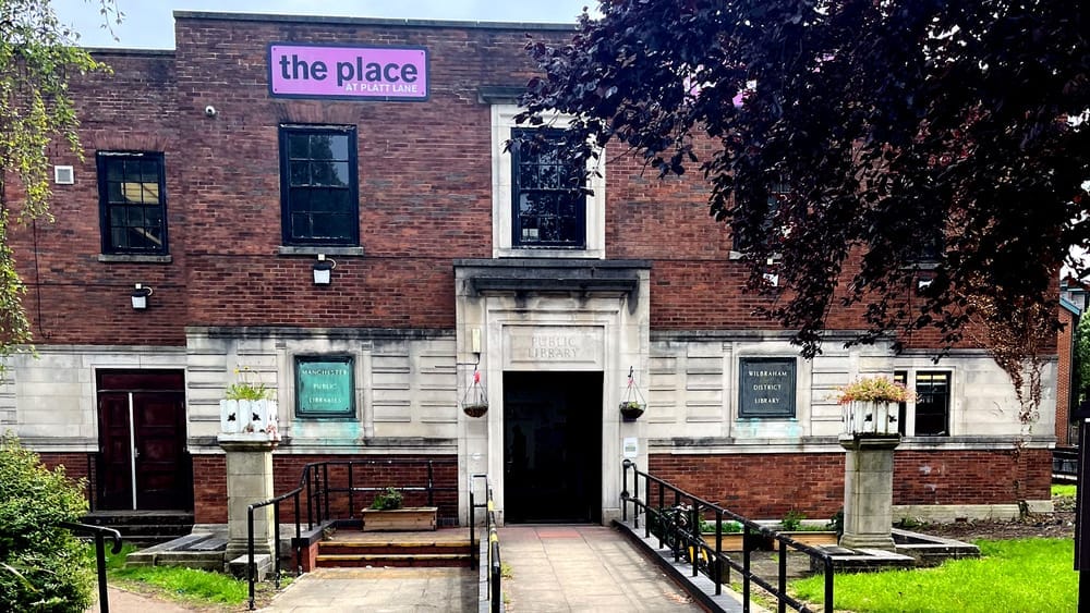 Handsome bronze plaques to either side of the wide door identify the Wilbraham District Library, Manchester; in the foreground, shade trees; at the top of the facade a lilac sign proclaims "The Place at Platt Lane"