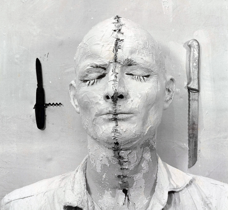 Self-portrait photograph of Günter Brus, thickly painted with makeup as if his face and head had been sewn up, alongside an open corkscrew and a serrated knife 