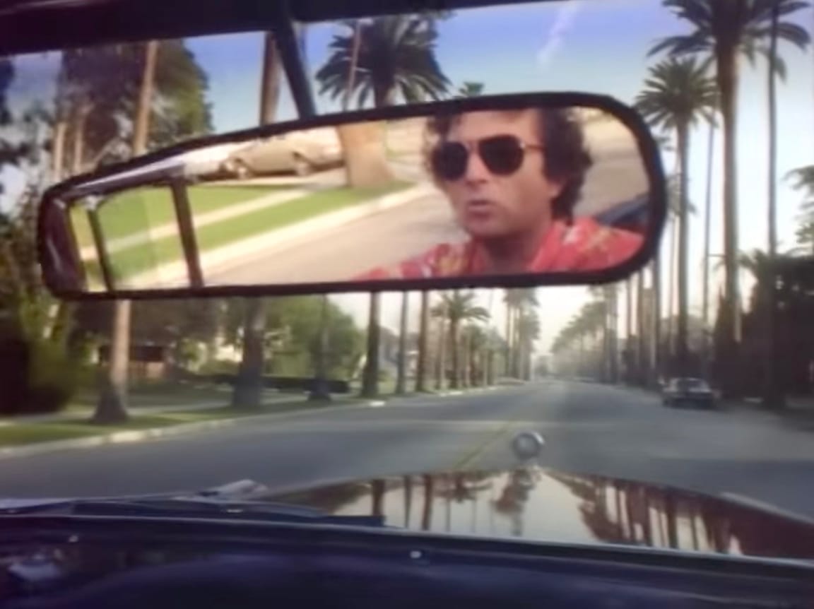 Randy Newman captured in rearview mirror of the car he's driving down a palm tree-lined street in the "I Love LA" video. 