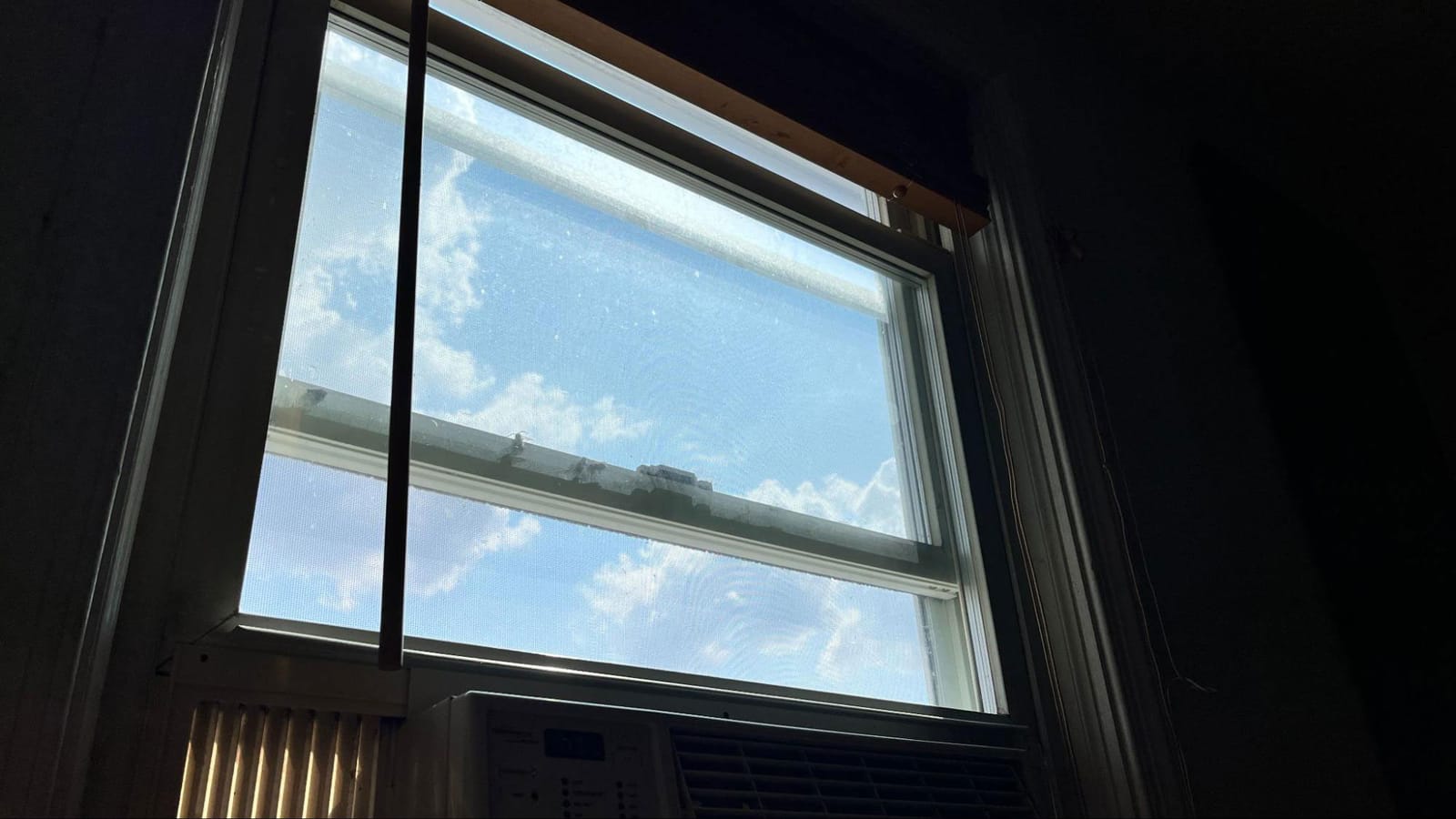 A window in a darkened room; outside, blue sky and scattered white clouds