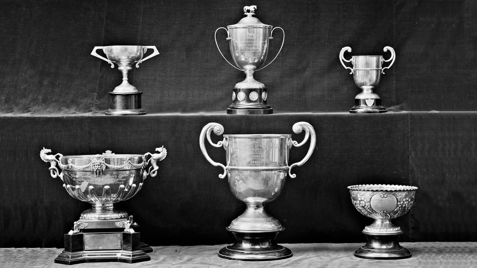 Black and white photo of six ornate silver trophies photographed against velvet