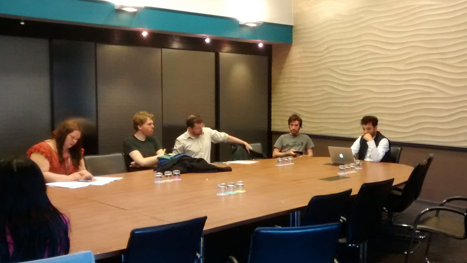 A boardroom with six people seated around a very large wooden table in a well-lit, rather featureless room